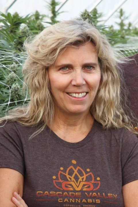 Theresa is smiling and wearing a Cascade Valley Cannabis shirt. She is in a greenhouse full of cannabis.